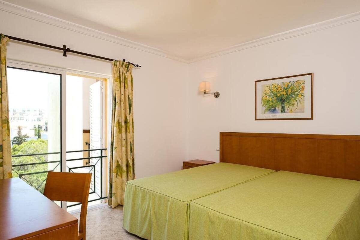 A hotel room with a bed, a desk, a chair, a nightstand, a lamp, and a window with a view of greenery.