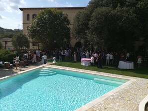 Ein Poolbereich mit einer terrasse aus Steinplatten, einer großen Menschenmenge und einem Gebäude mit Bogenfenstern im Hintergrund.