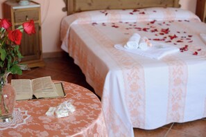 Ein Schlafzimmer mit einem mit Rosenblättern bezogenen Bett, einem Nachttisch mit einem Buch und einer Rosenvase sowie einem Holznachttisch.