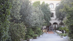 A courtyard with potted plants, a building with arched windows, and a few people sitting at tables. - Hotel Funtana Noa (Villanovaforru)