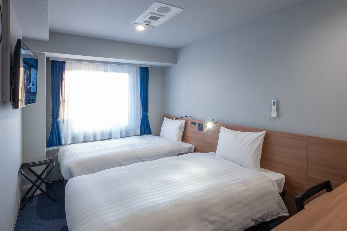 A hotel room with two beds, a desk, a chair, a TV, and a window with curtains.
