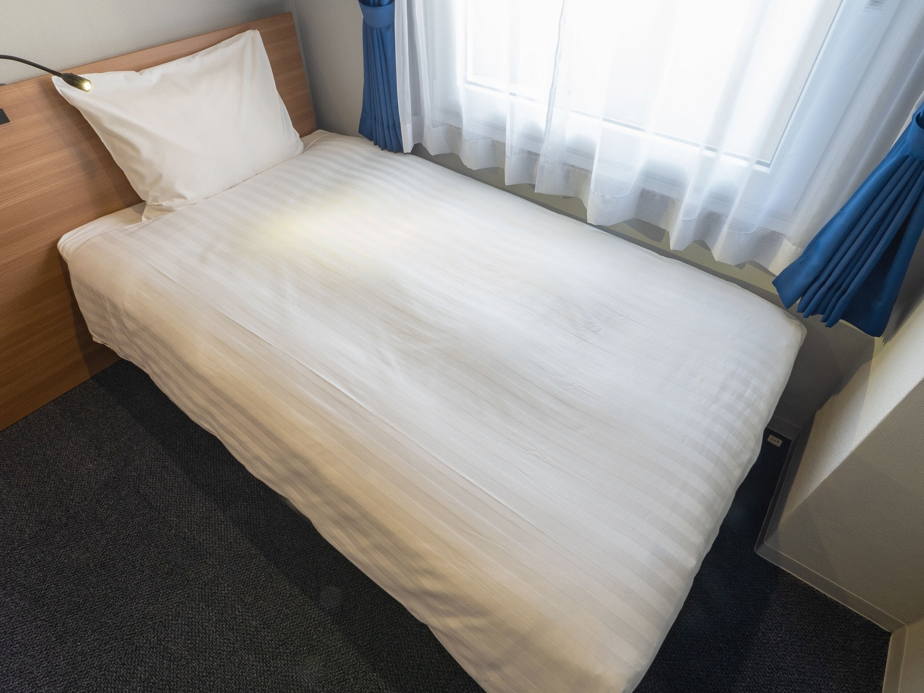 A single bed with a wooden headboard, a bedside table, and a window with sheer curtains.