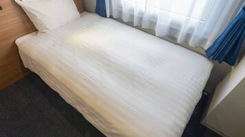 A single bed with a wooden headboard, a bedside table, and a window with sheer curtains.