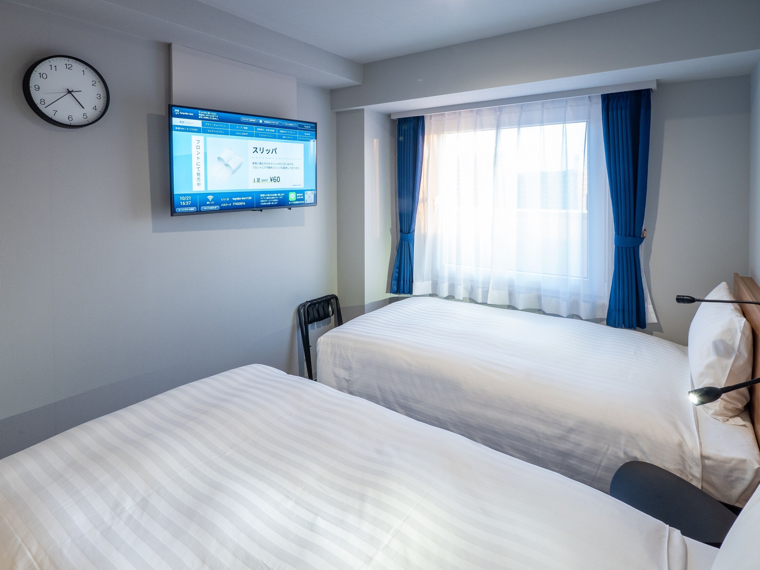 Habitación de hotel con dos camas, un reloj de pared, una pantalla digital y una ventana con cortinas.