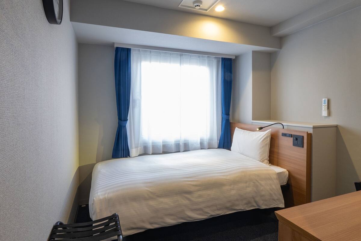 A hotel room with a bed, a desk, a chair, a window with curtains, and a wall-mounted control panel.