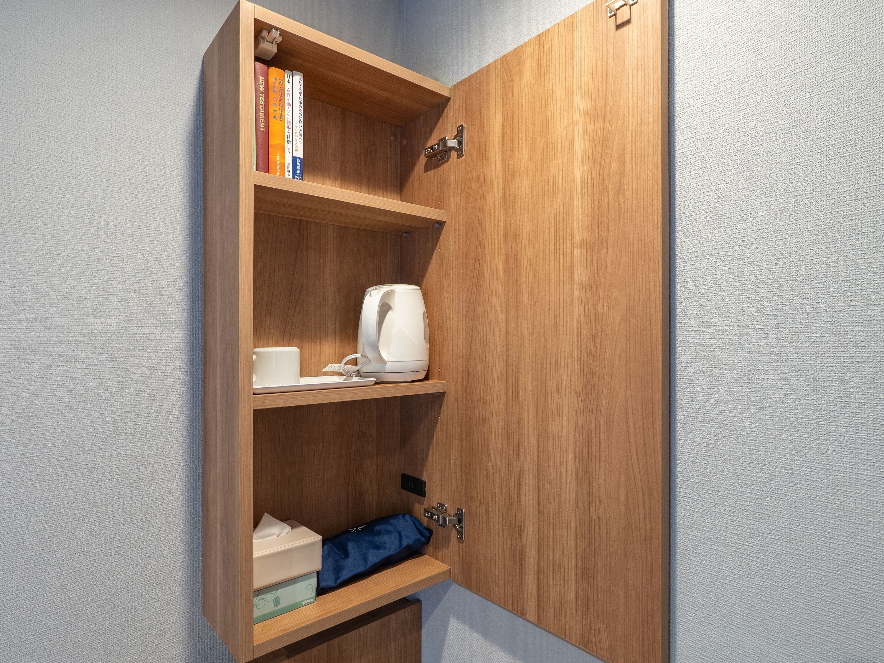 A wooden cabinet with shelves containing books, a kettle, a cup, and a tissue box.
