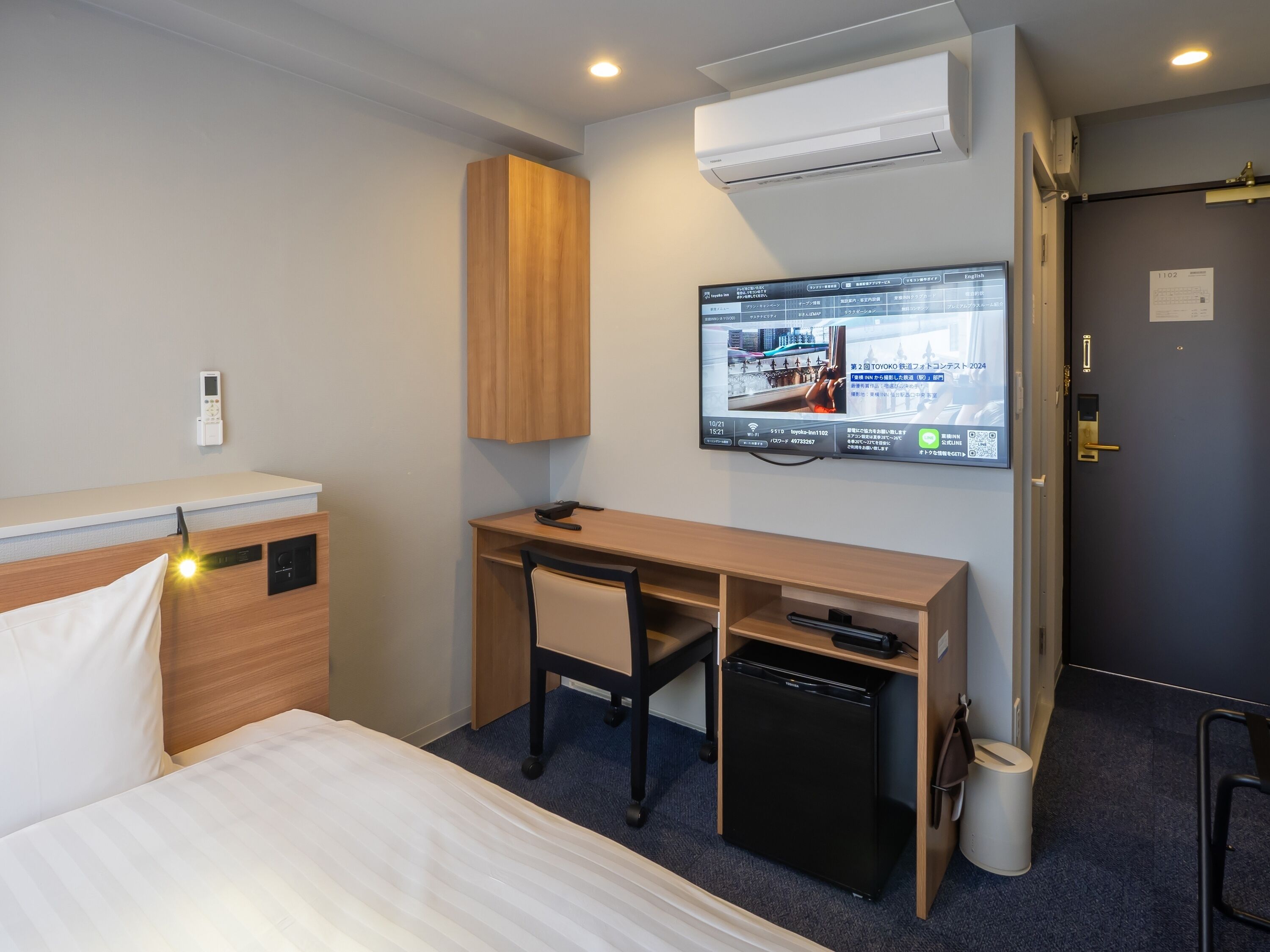 A hotel room with a bed, desk, chair, TV, air conditioning unit, and a small refrigerator.