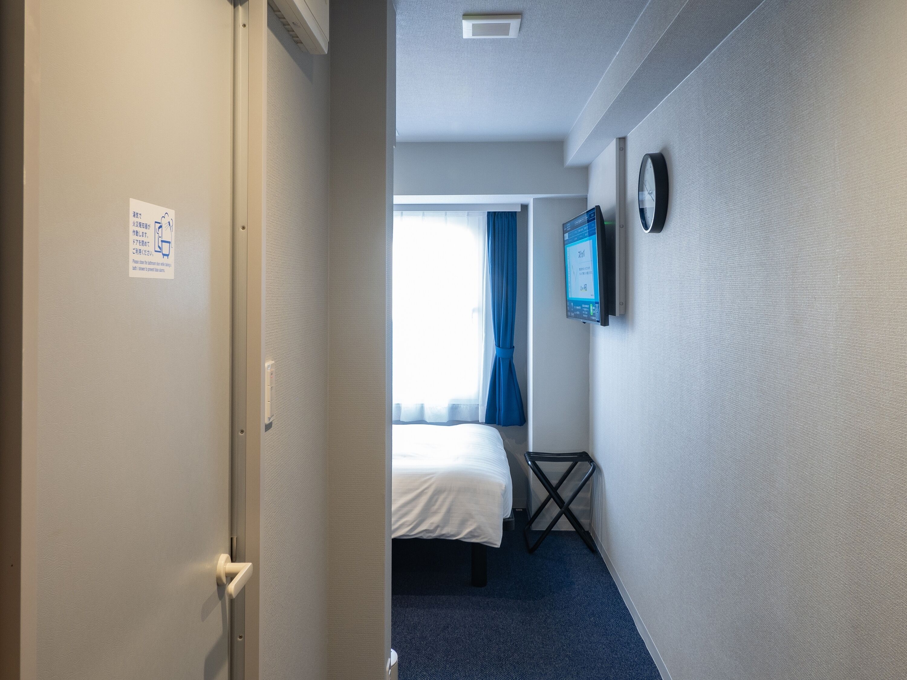 Une chambre d’hôtel avec un lit, un téléviseur fixé au mur, une horloge et une porte avec une pancarte.