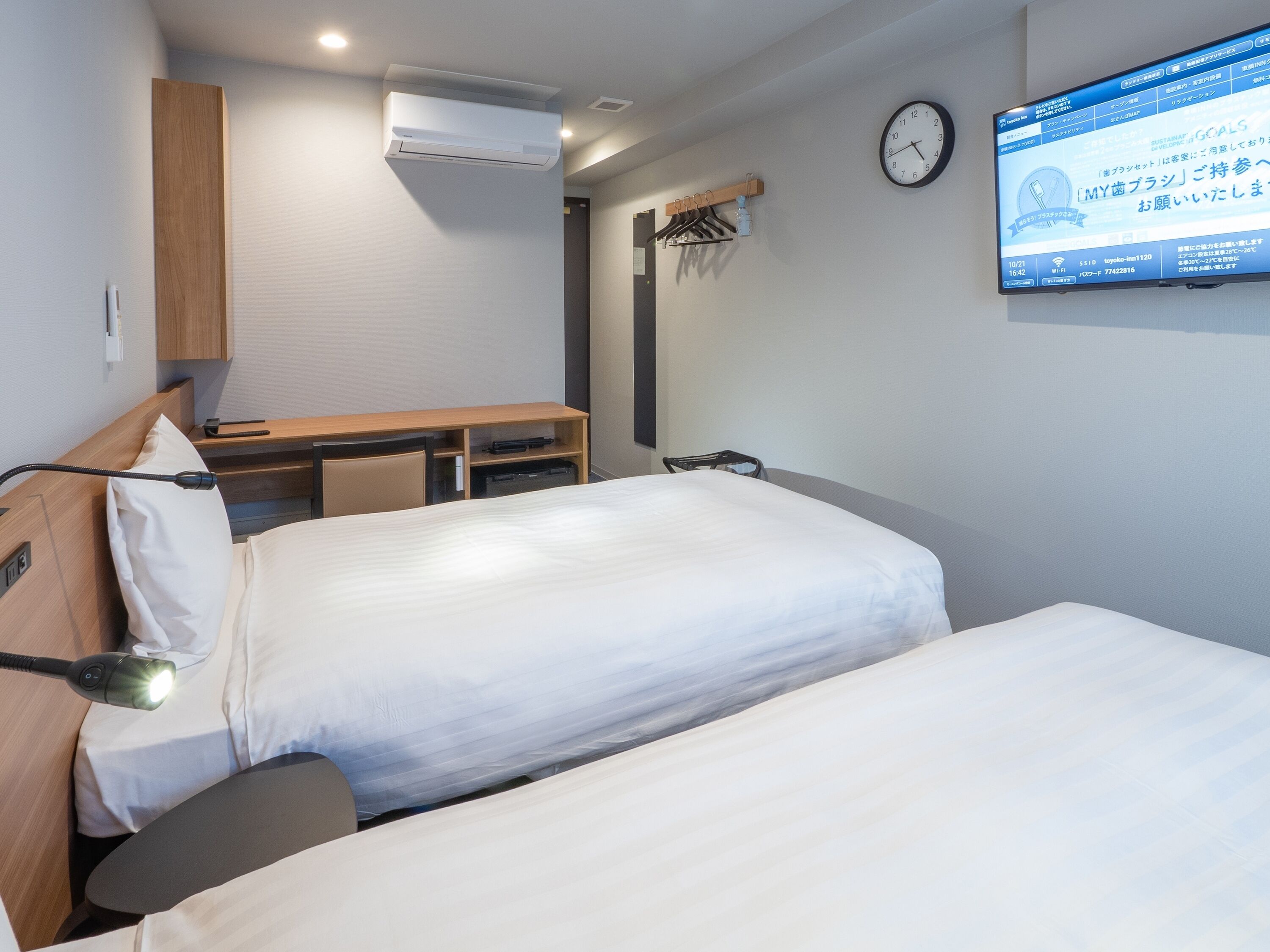 Une chambre d’hôtel avec deux lits, un bureau, un appareil de climatisation, une horloge et un téléviseur affichant du texte en japonais.