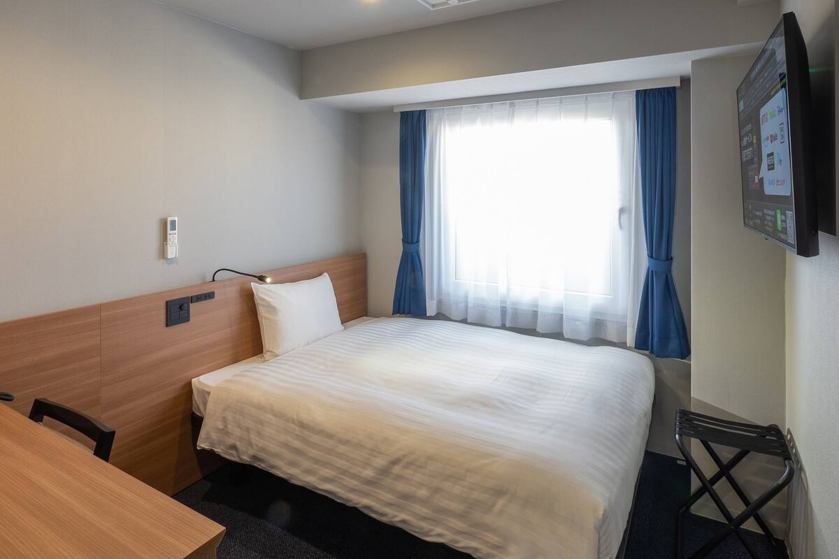 A hotel room with a bed, a desk, a chair, a window with curtains, and a wall-mounted control panel.