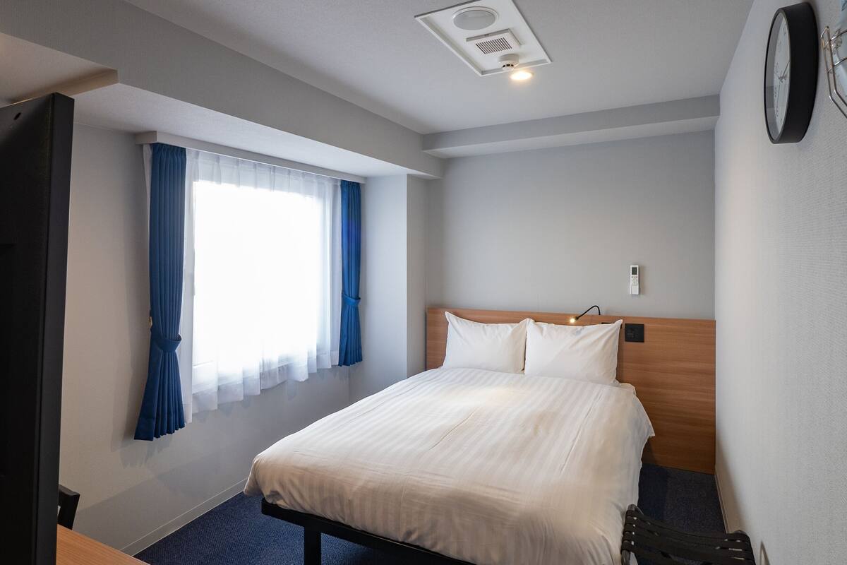 A hotel room with a bed, a desk, a chair, a window with curtains, a wall clock, and a ceiling vent.