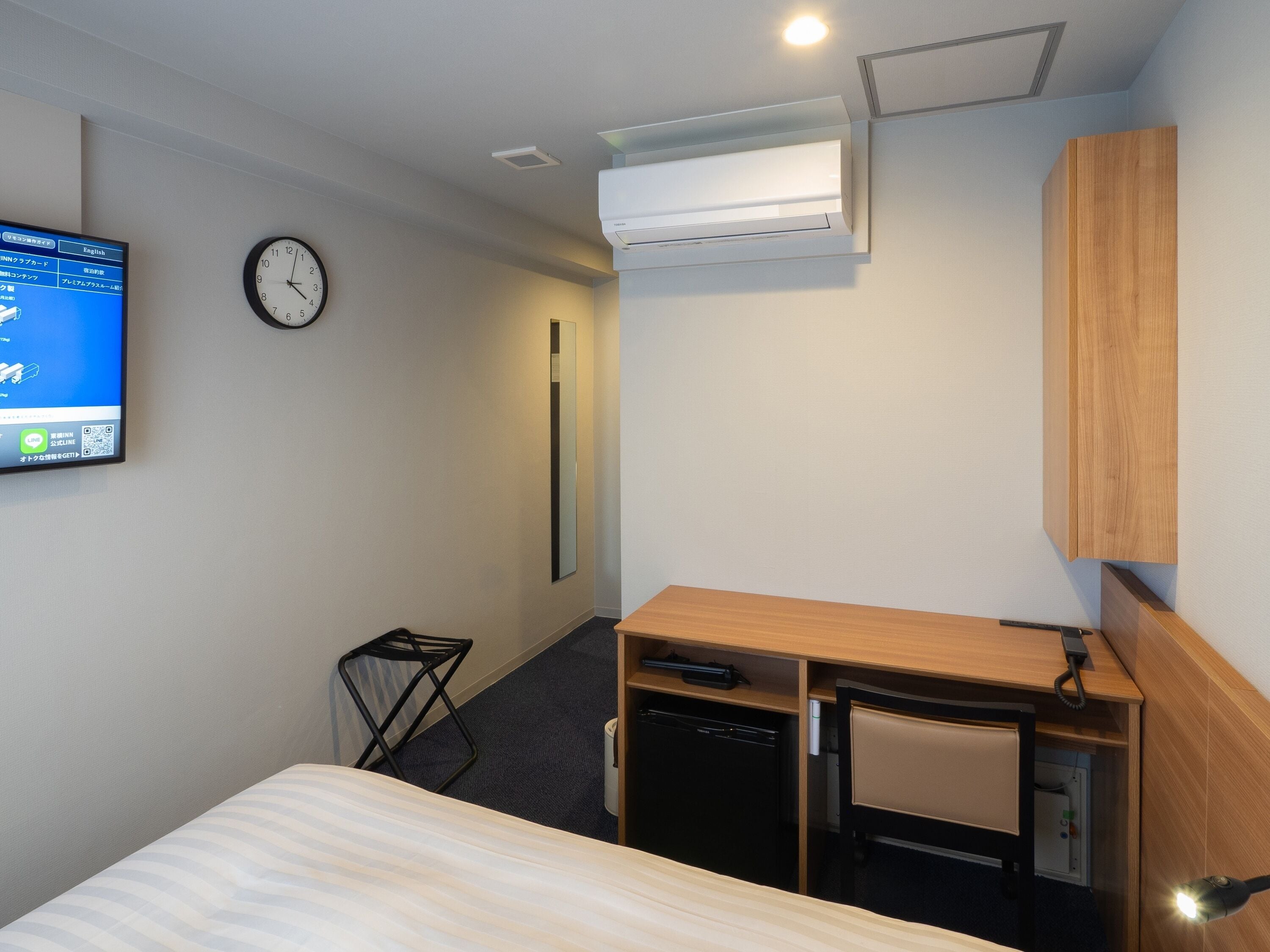 A hotel room with a bed, a desk with a chair, a small refrigerator, a wall-mounted clock, and a TV displaying a map.