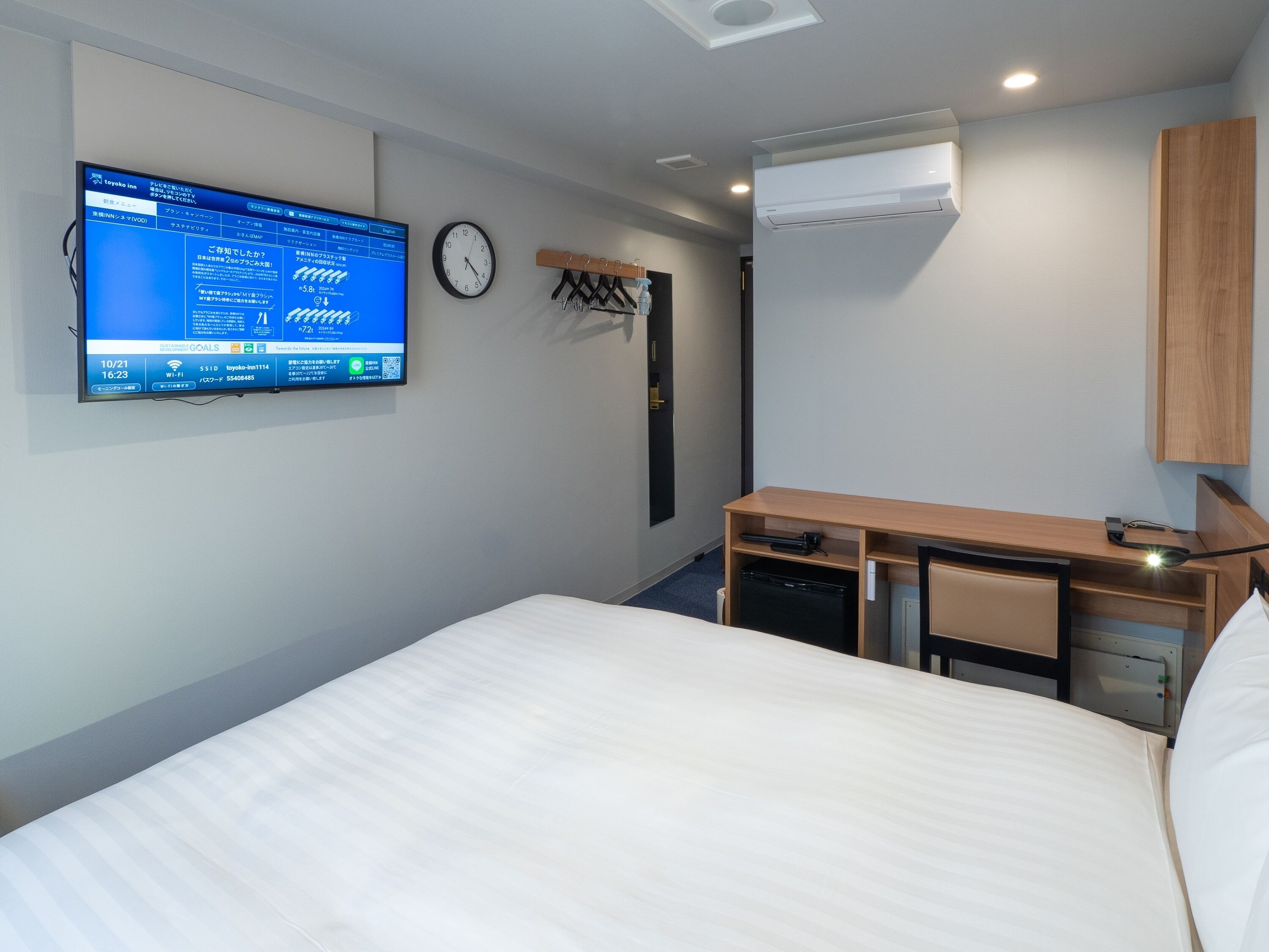 A hotel room with a bed, a desk with a computer, a clock, and an air conditioning unit.