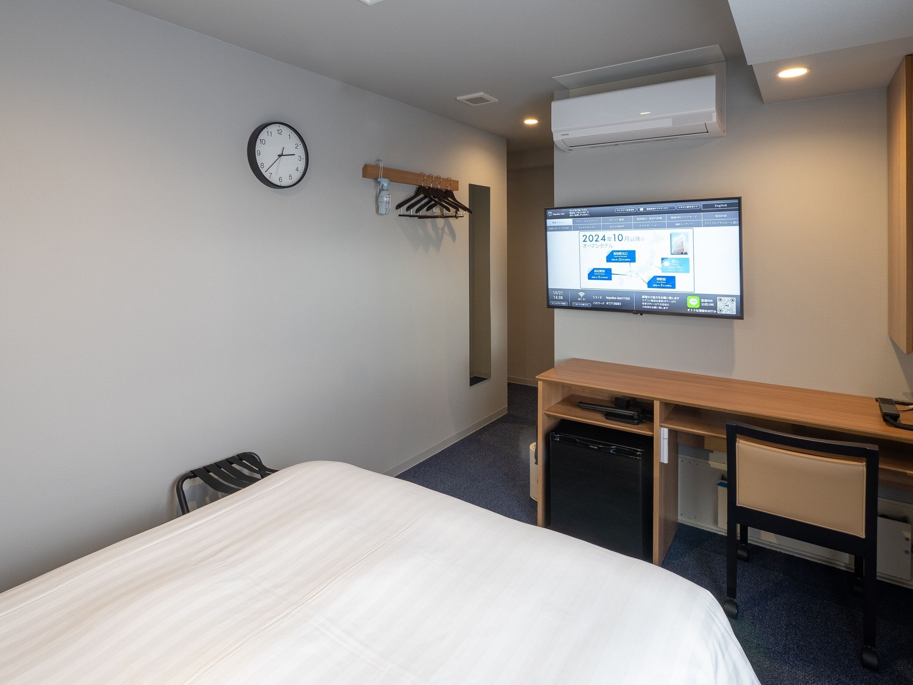 A hotel room with a bed, a desk with a chair, a clock, and a TV displaying a map.