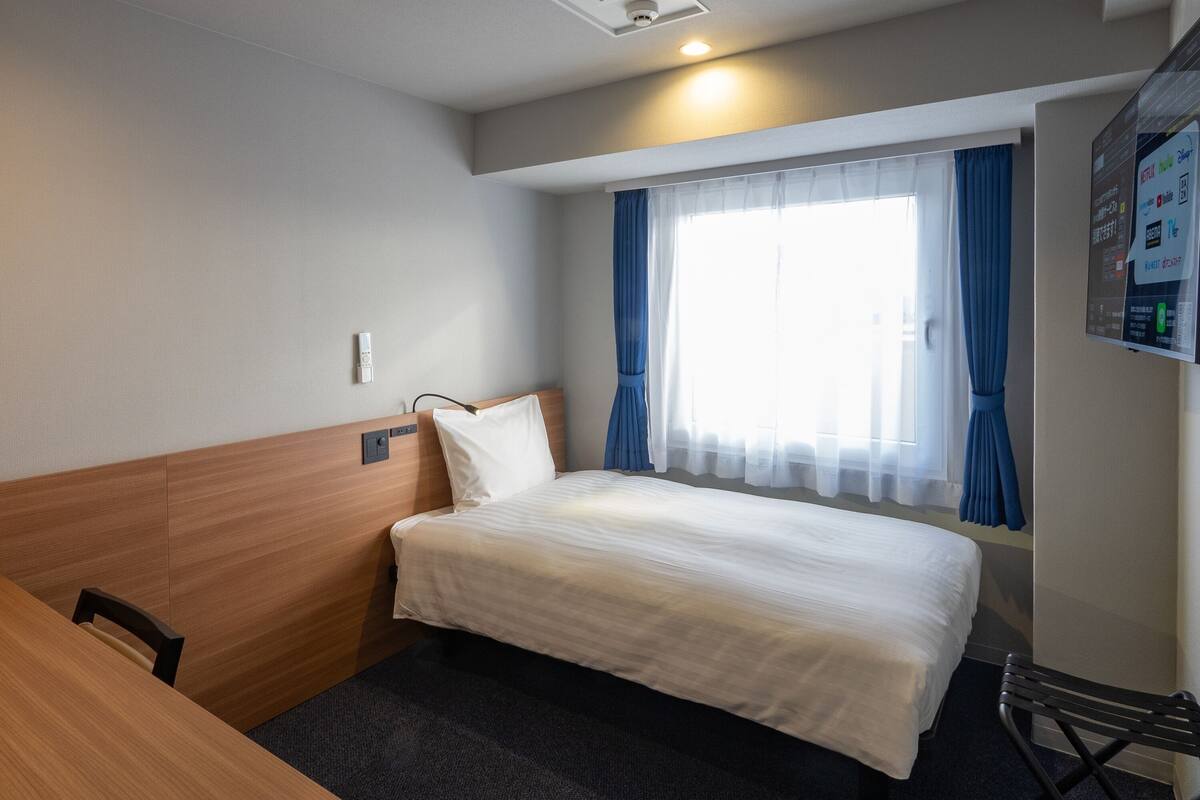 A hotel room with a bed, a desk, a chair, a window with curtains, and a wall-mounted control panel.