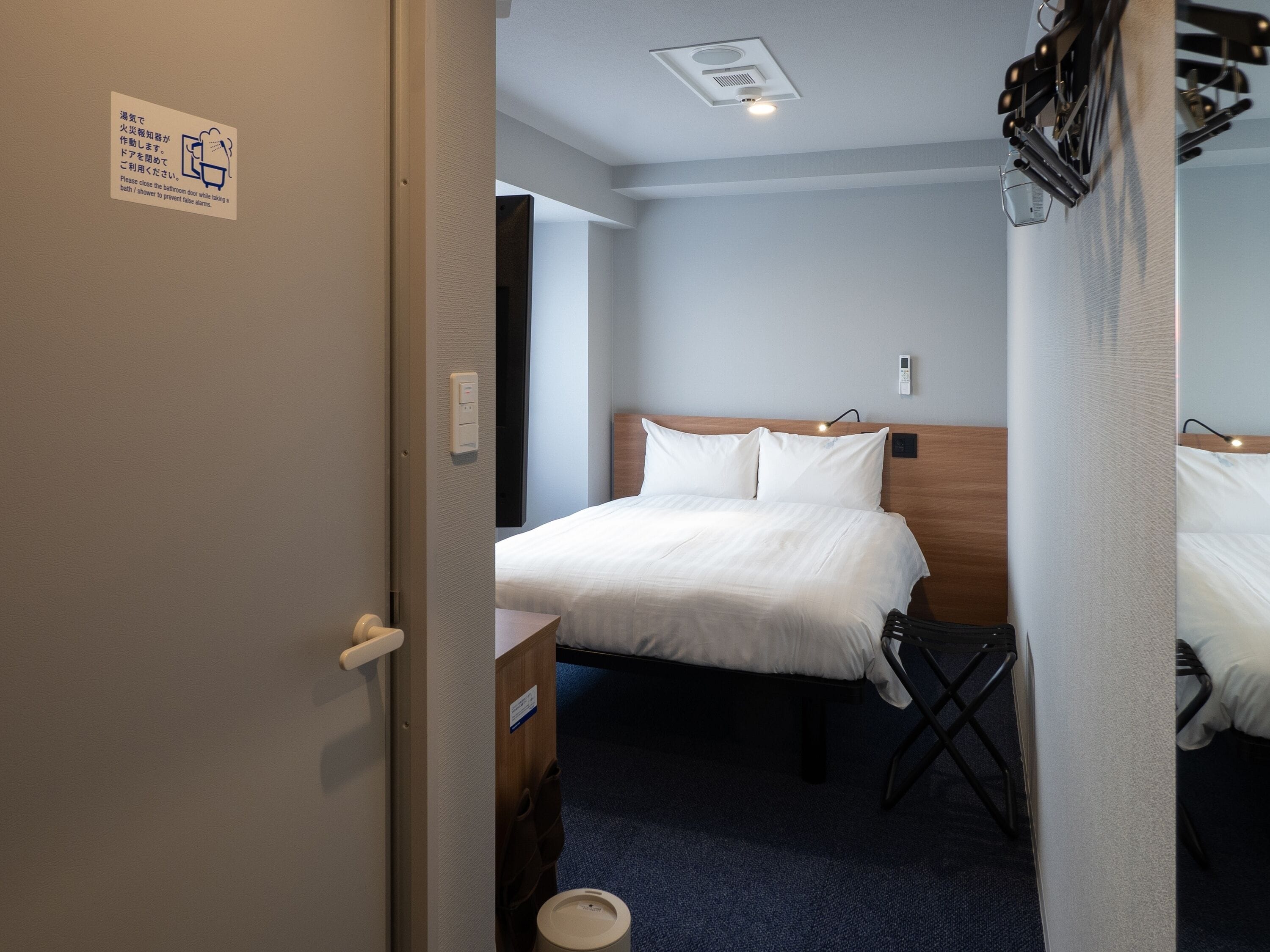 A hotel room with a bed, a bedside table, a wall-mounted light, and a ceiling vent.
