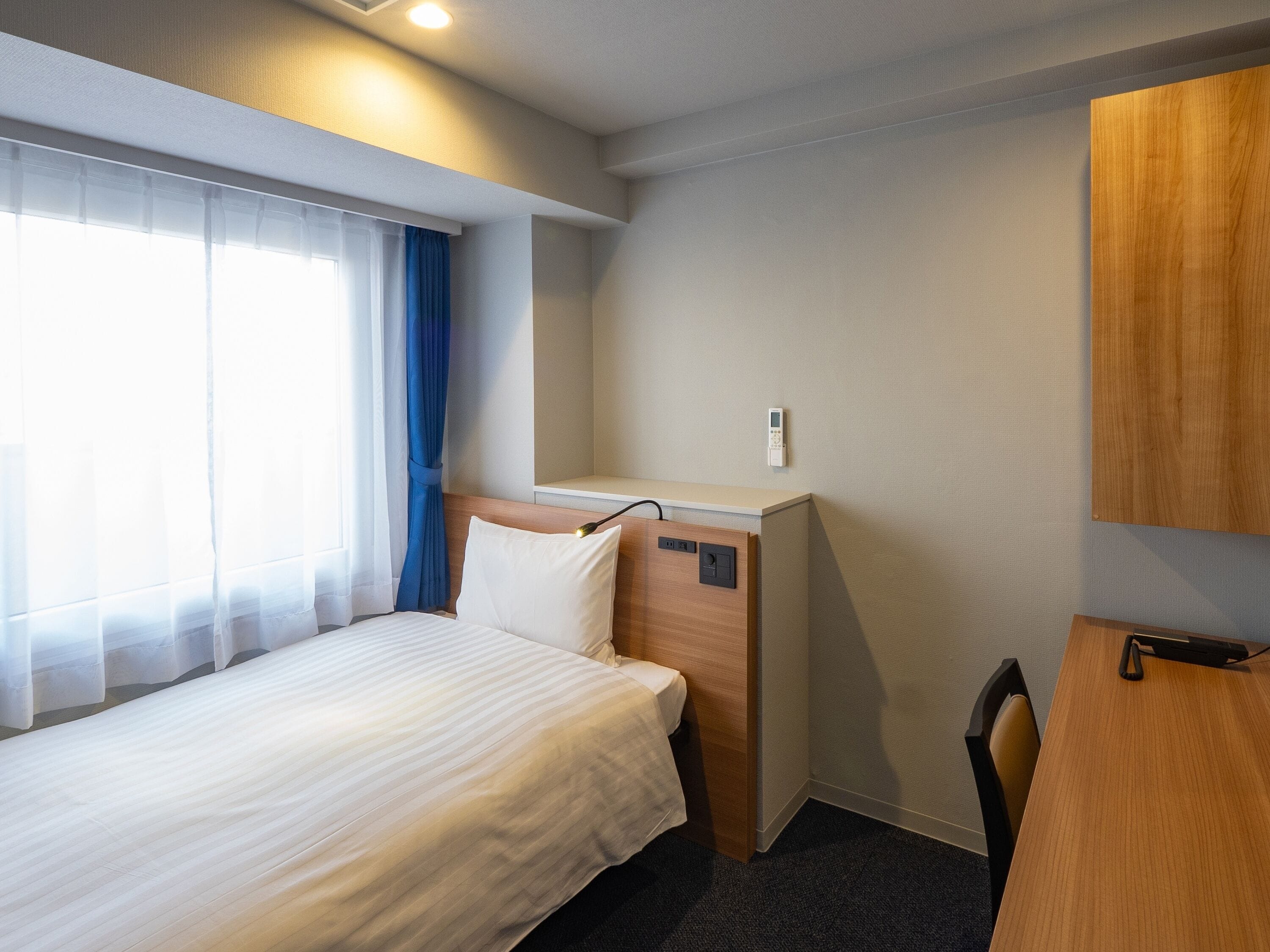 A hotel room with a bed, a desk, a chair, a window with curtains, and a wall-mounted control panel.