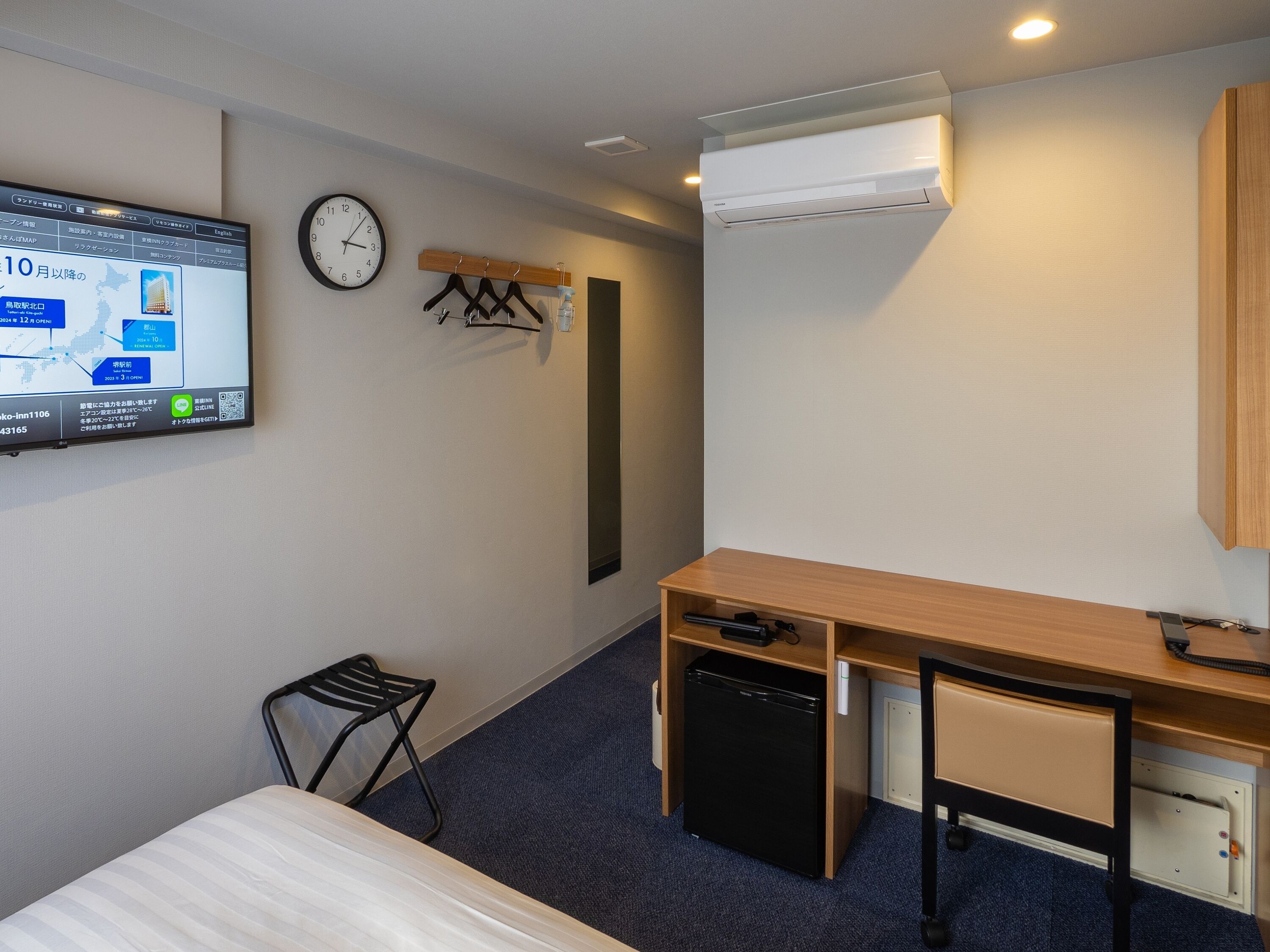 A hotel room with a bed, a desk with a computer, a small refrigerator, a chair, a clock, and a wall-mounted TV displaying a map.