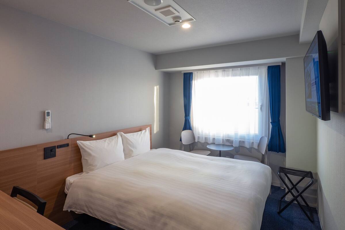A hotel room with a bed, bedside table, chair, TV, and a window with curtains.