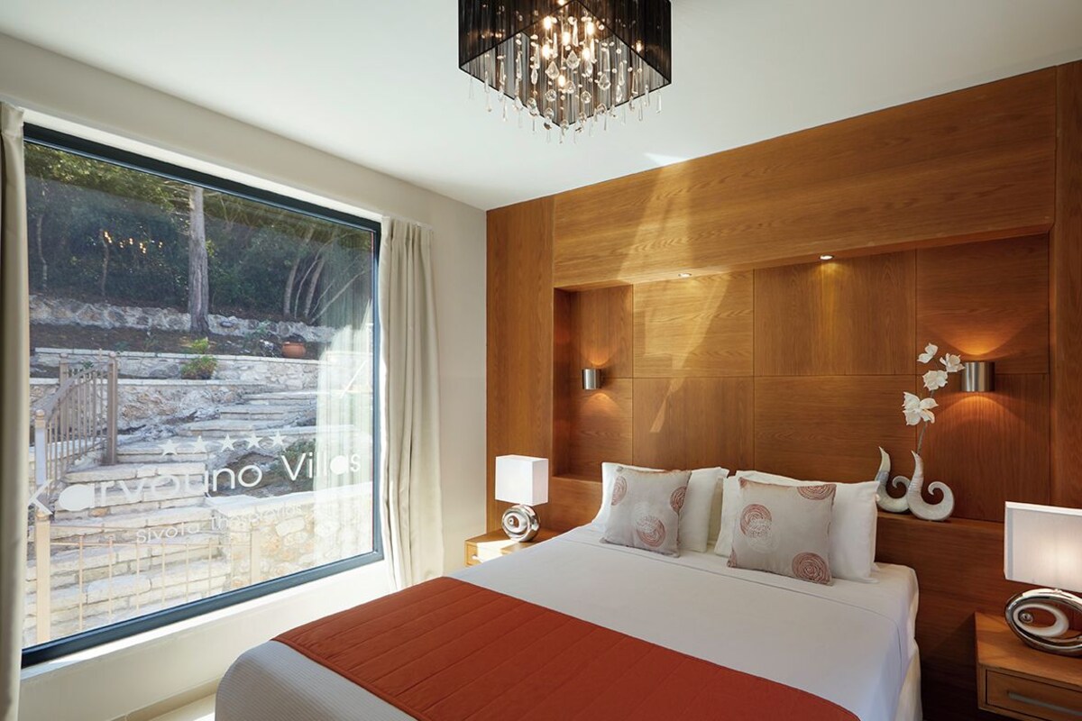 Ein modernes Hotelzimmer mit einem großen Bett, einem Holzkopfteil, einer Kronleuchter und Blick auf eine Steinmauer und Grünflächen.