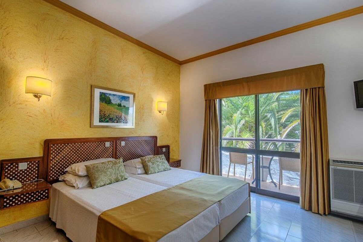 A hotel room with a large bed, two bedside lamps, a painting on the wall, a window with curtains, and a view of palm trees.