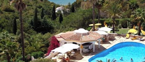 Uma área de piscina com espreguiçadeiras e guarda-sóis, cercada por vegetação e com vista para um corpo d'água.