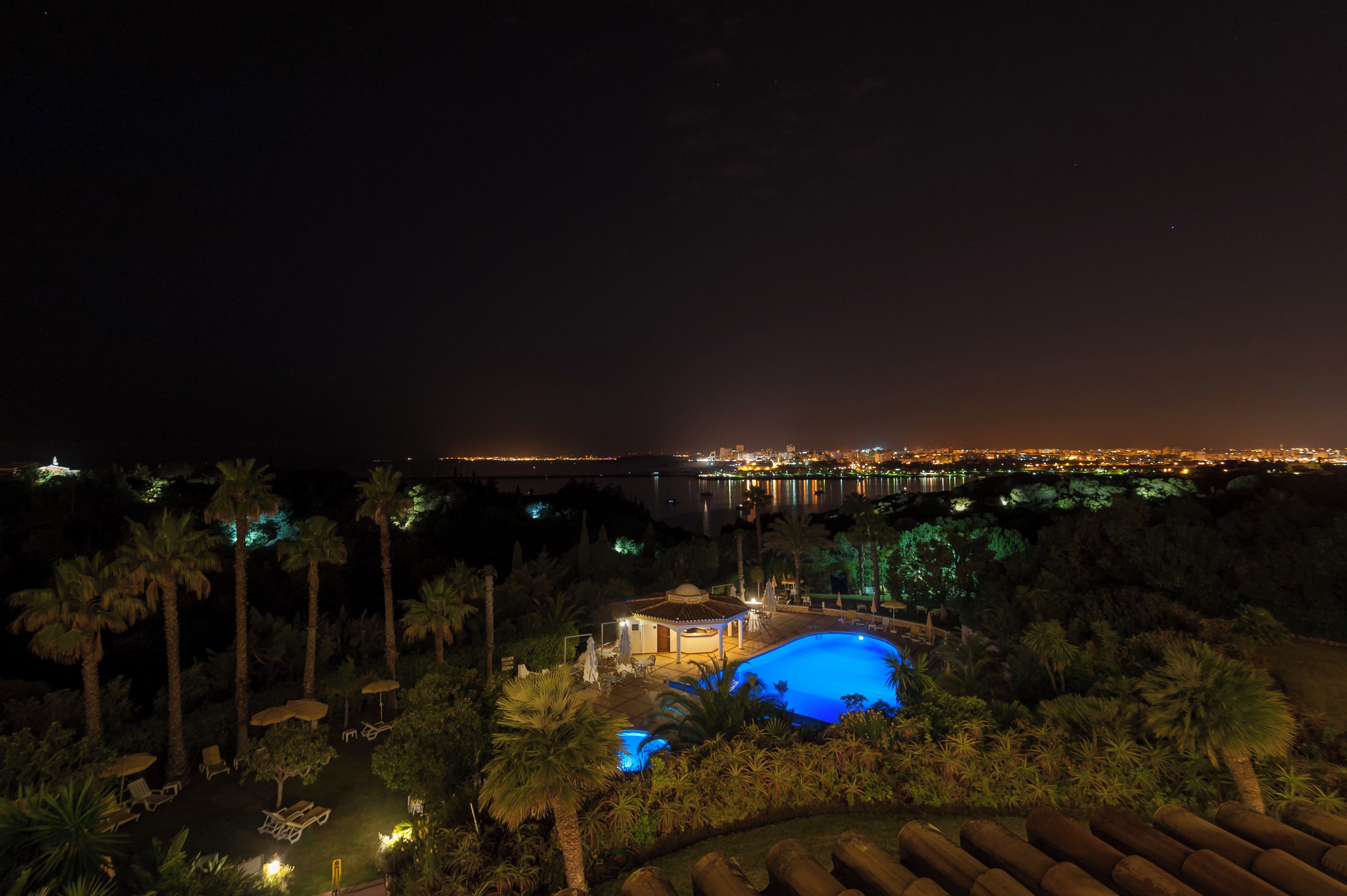 A nighttime view of a resort with a pool, palm trees, and a cityscape in the background.