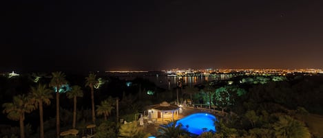 Vista nocturna de un resort con piscina, palmeras y un paisaje urbano al fondo.