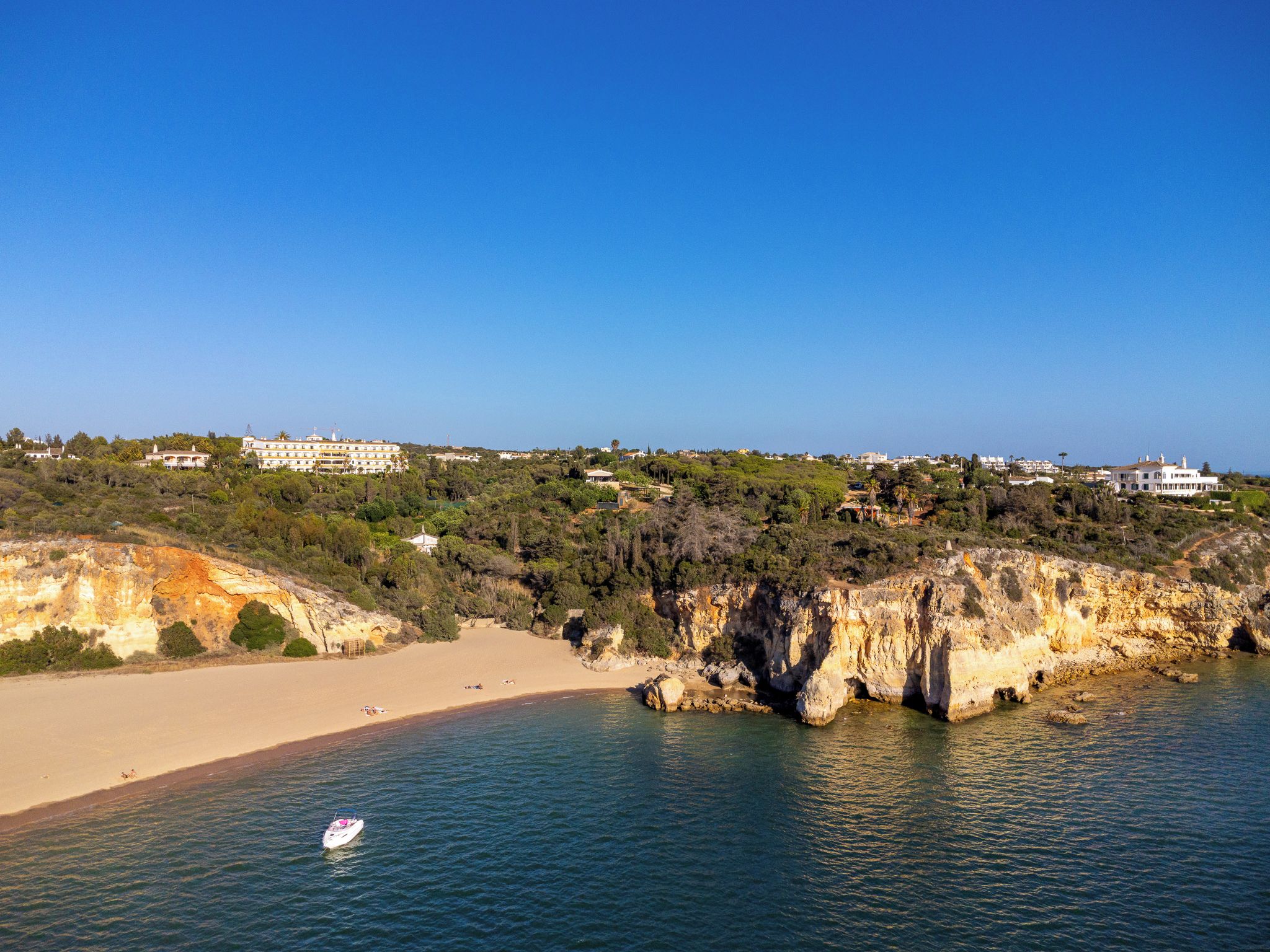 Uma área costeira com uma praia de areia, falésias rochosas e edifícios ao fundo.