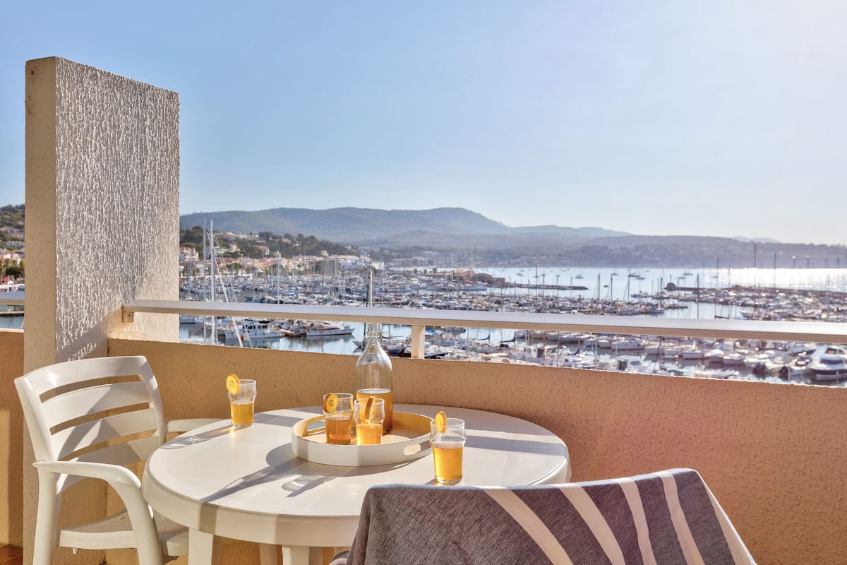 A balcony with a table set for two, overlooking a marina with boats and distant hills.