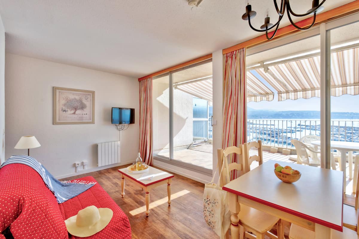 A room with a sofa, a dining table, and a TV. There is a window with a view of the sea.