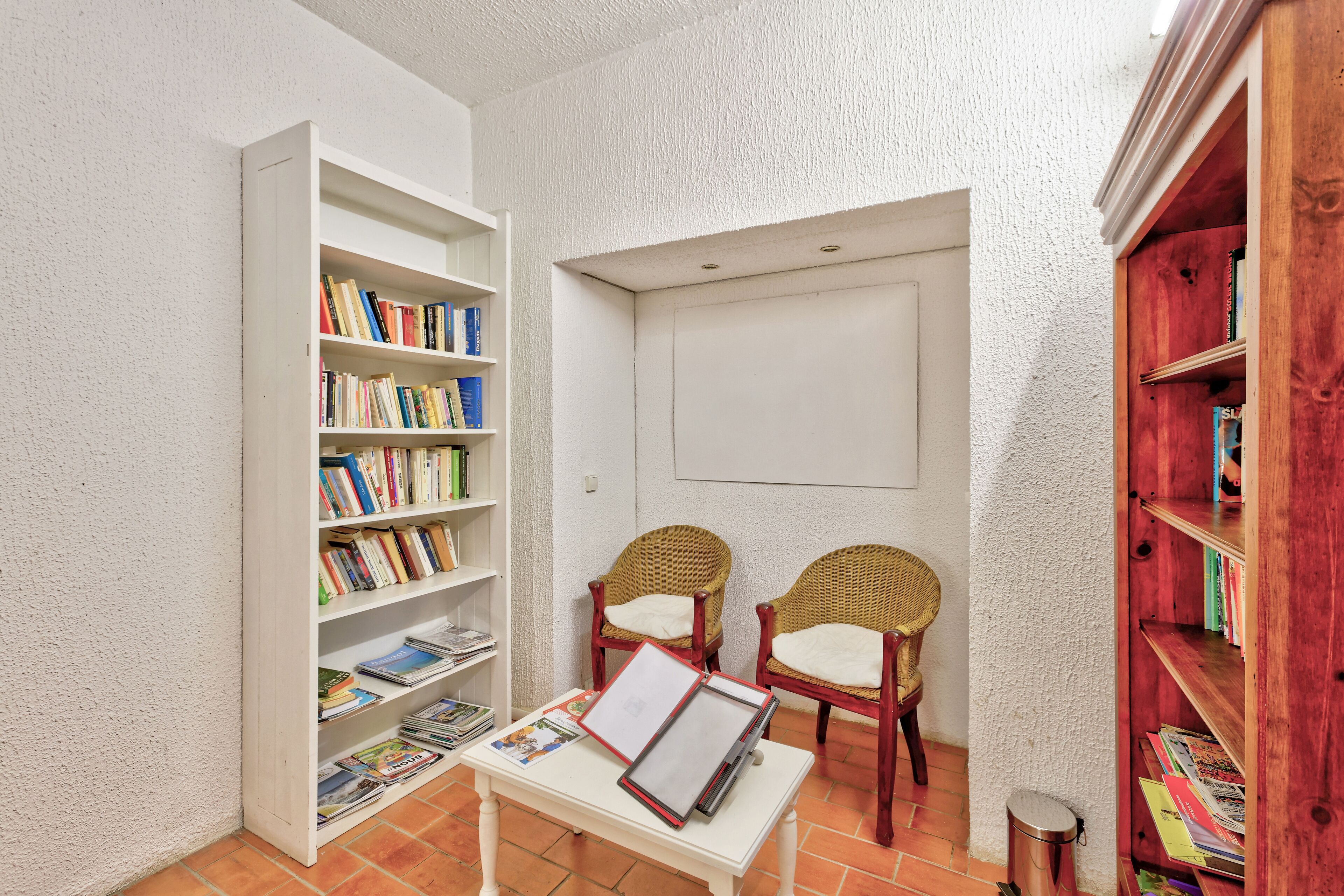 A reading area with a white bookshelf, two wicker chairs, a small table with magazines, and a red bookshelf.