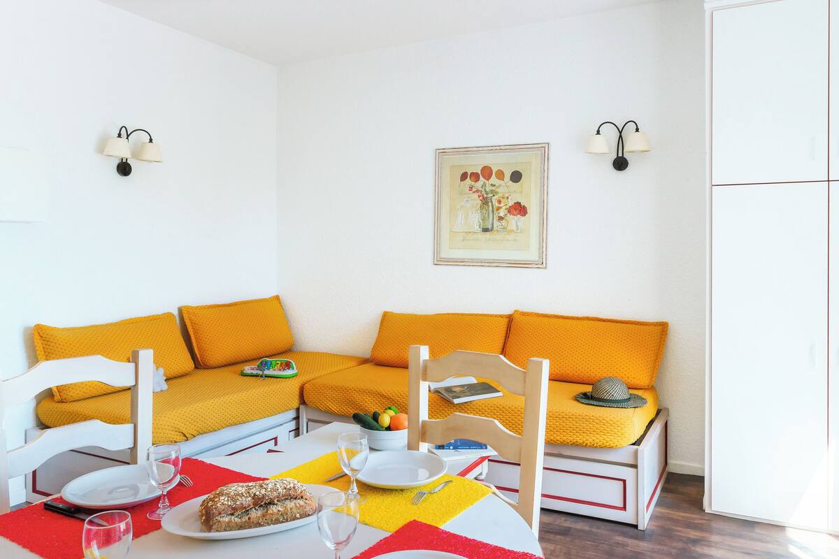 A dining area with a table set for two, a sofa with yellow cushions, and a framed picture on the wall.