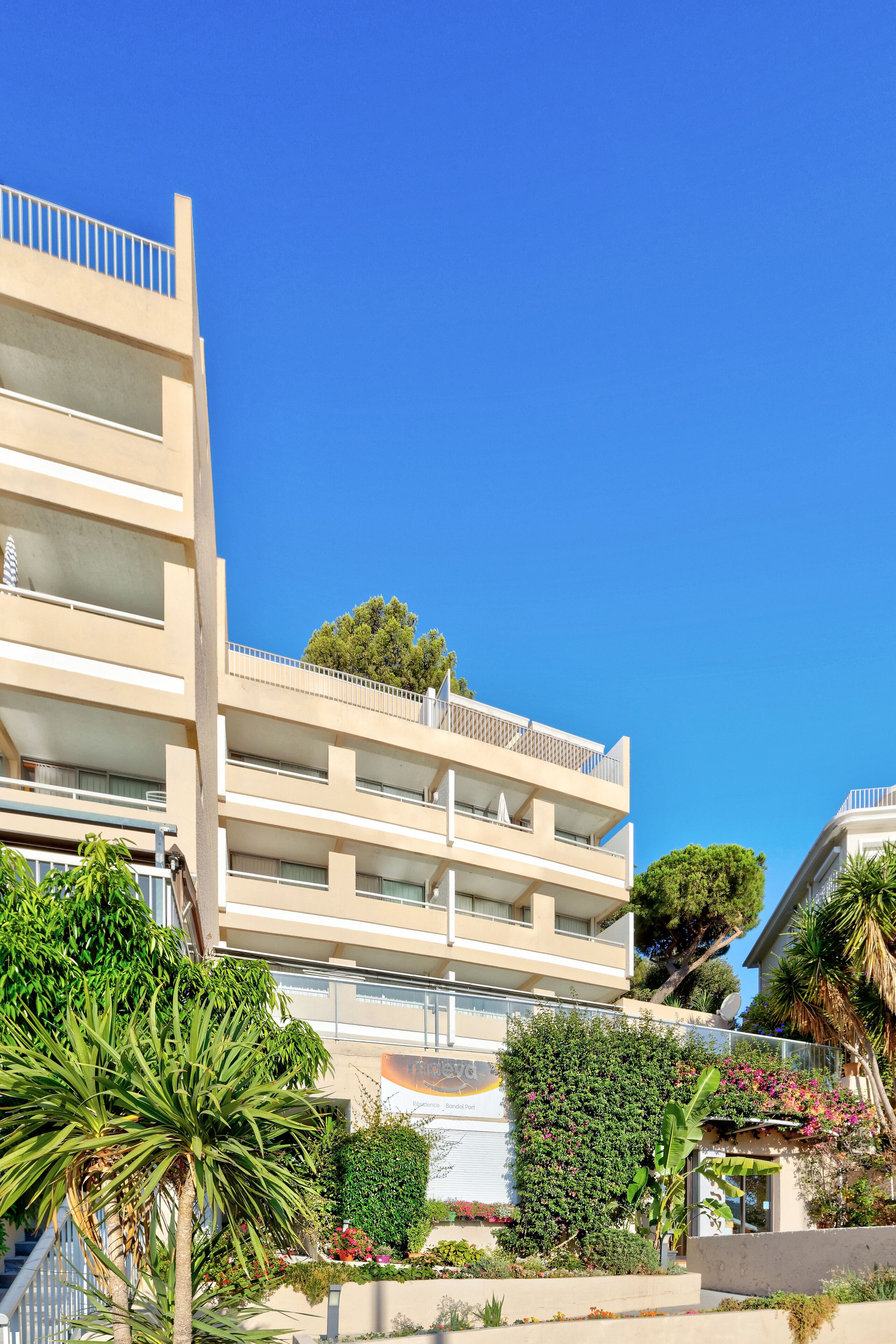 Un hôtel à plusieurs étages doté de balcons, d’un jardin agrémenté de diverses plantes et surplombant un ciel d’un bleu limpide.