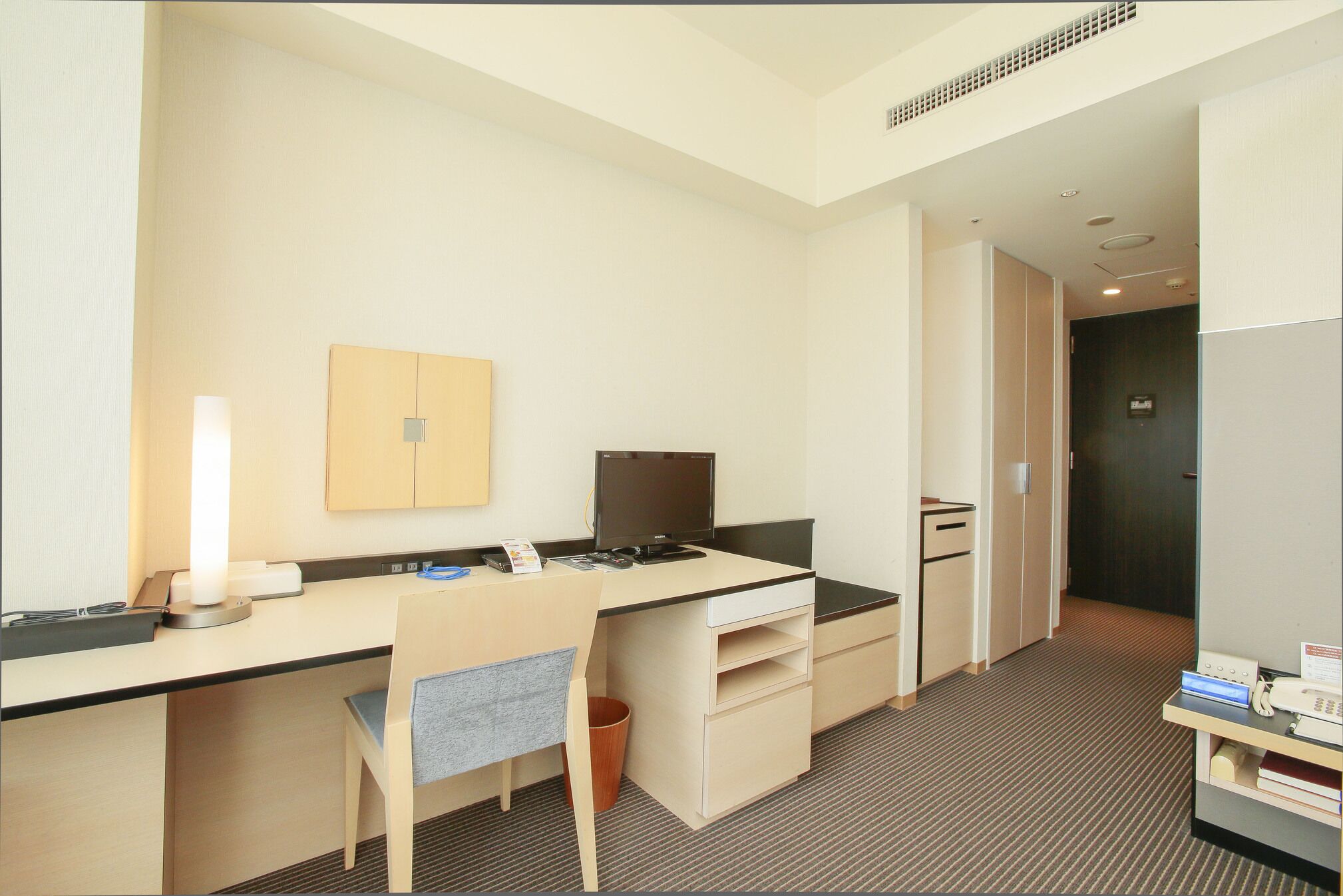 A hotel room with a desk, chair, computer, and a small trash bin.
