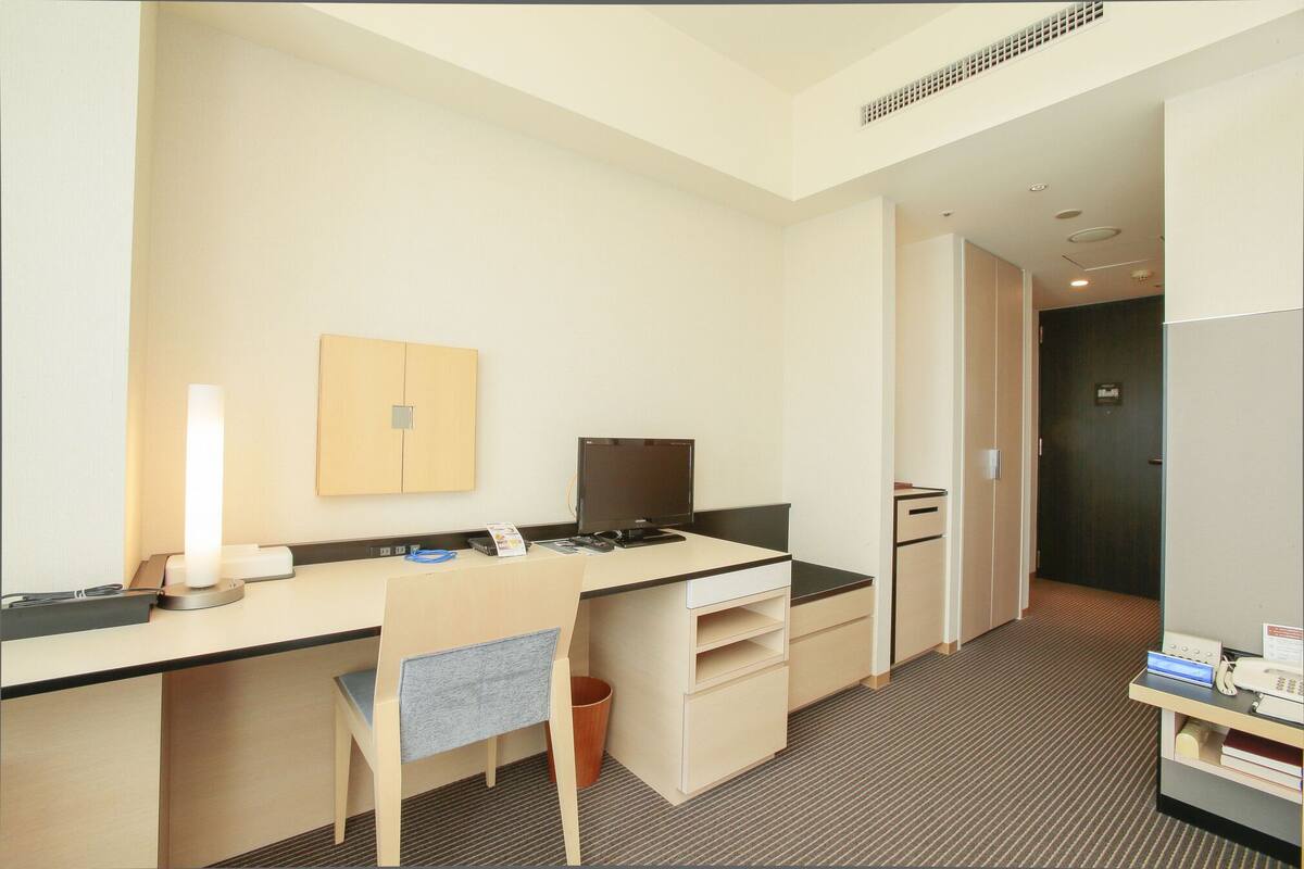 A hotel room with a desk, chair, computer, and a small trash bin.