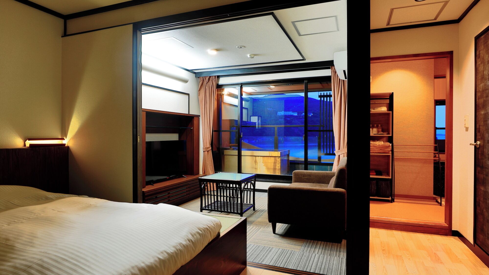 A hotel room with a bed, a desk, a chair, a TV, and a view of a cityscape through a large window.