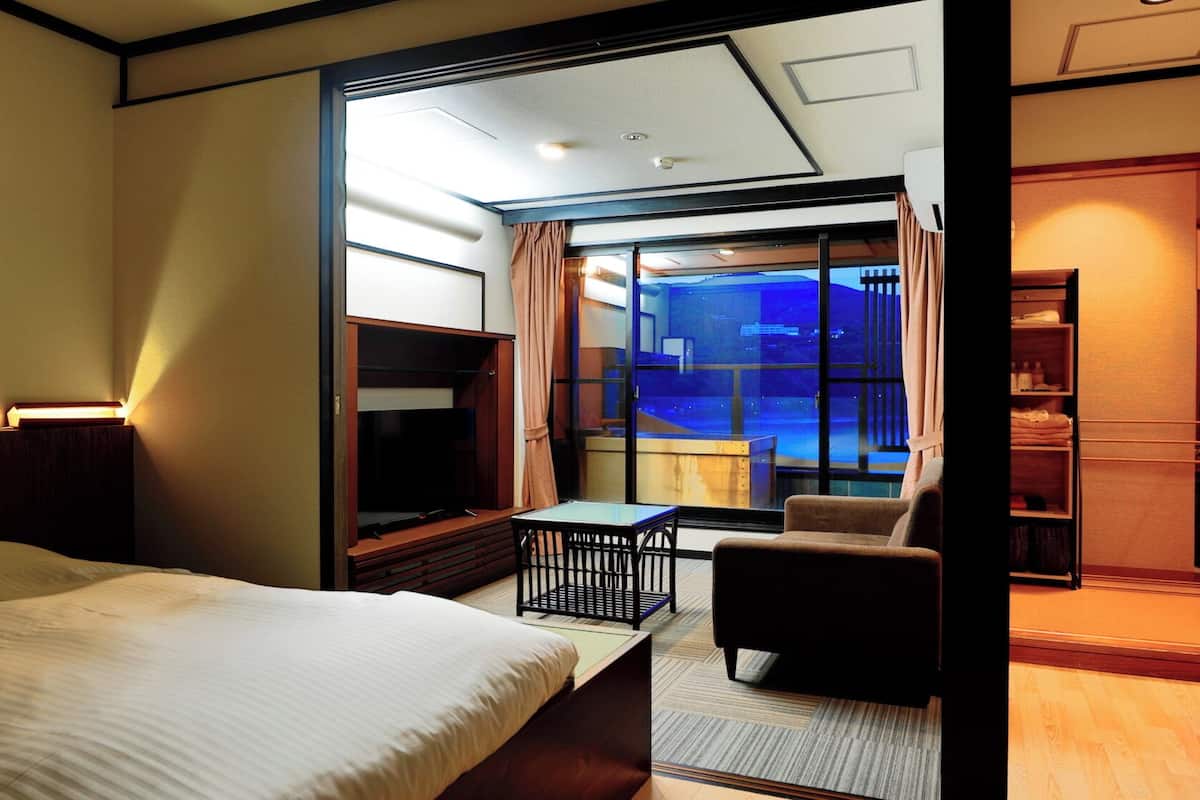 A hotel room with a bed, a desk, a chair, a TV, and a view of a cityscape through a large window.