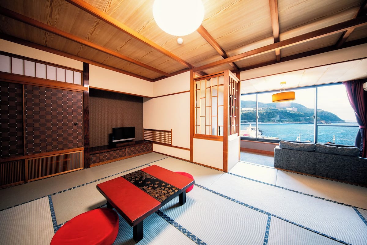 A traditional Japanese room with tatami flooring, a low table, and a sofa by a large window overlooking a body of water.