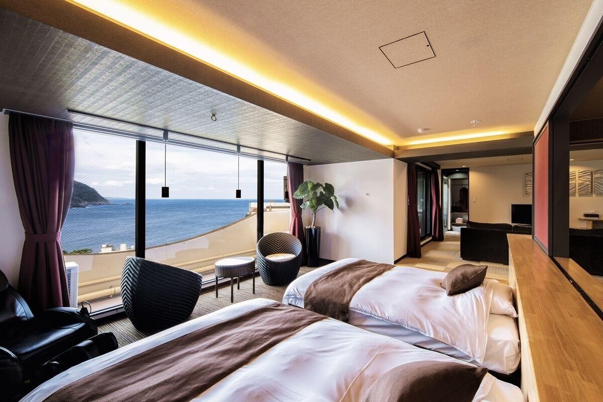 A modern hotel room with two beds, a seating area, a view of the sea, and a balcony.