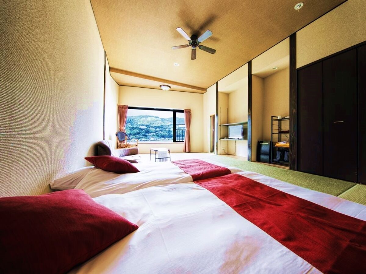 A hotel room with two beds, a ceiling fan, a large window, and a view of the outdoors.