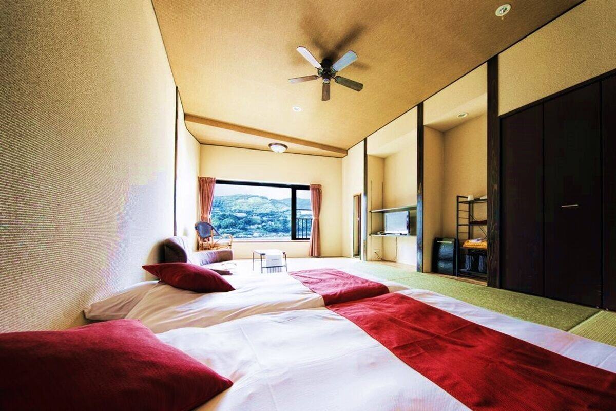 A hotel room with two beds, a ceiling fan, a large window, and a view of the outdoors.