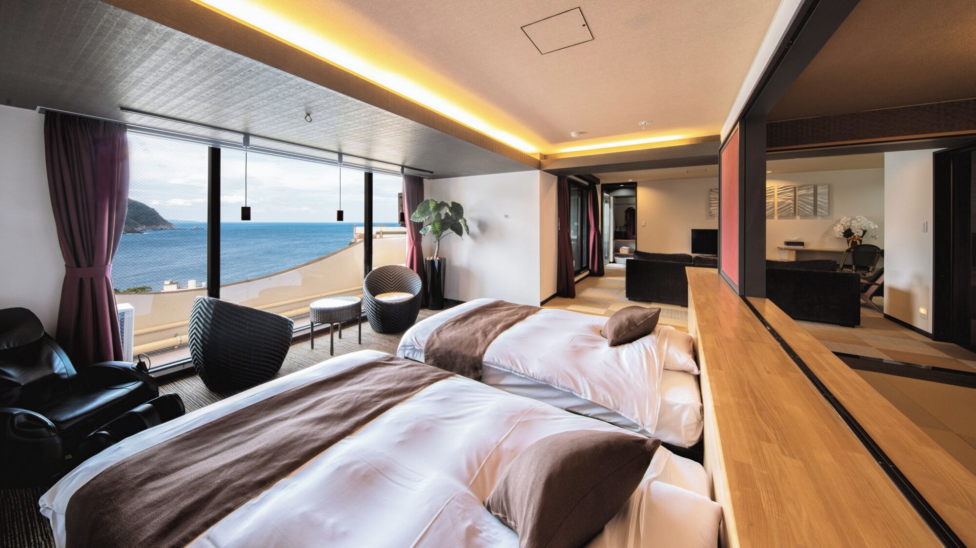 A modern hotel room with a large bed, a seating area, a desk, and a view of the sea.