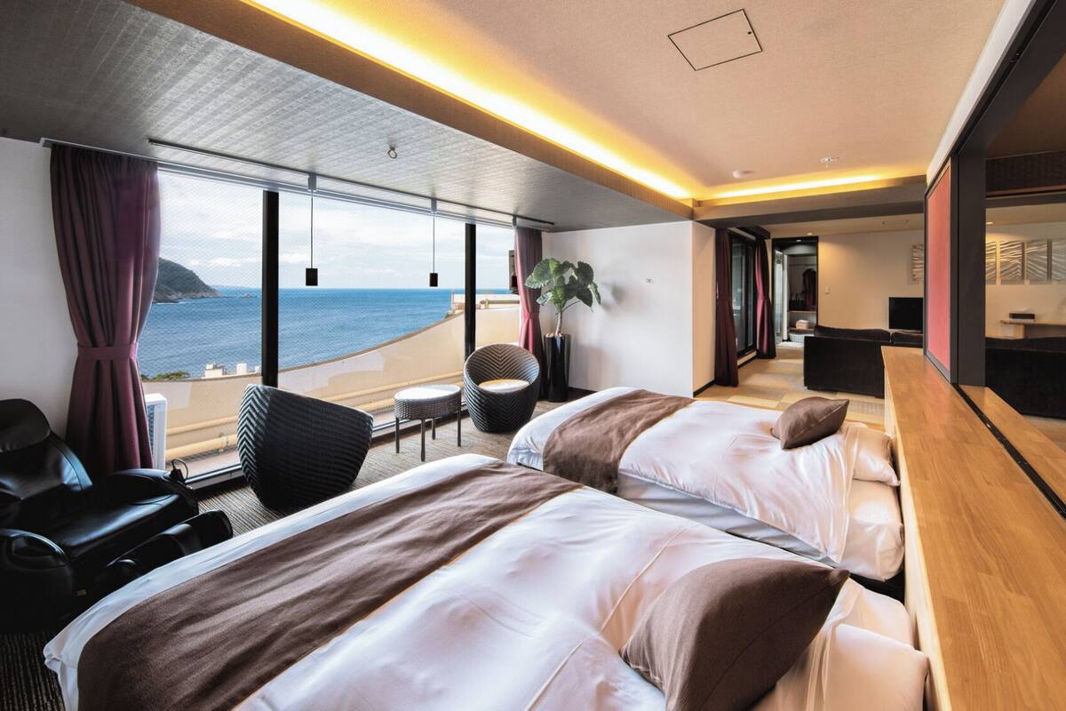 A modern hotel room with a large bed, a seating area, a desk, and a view of the sea.