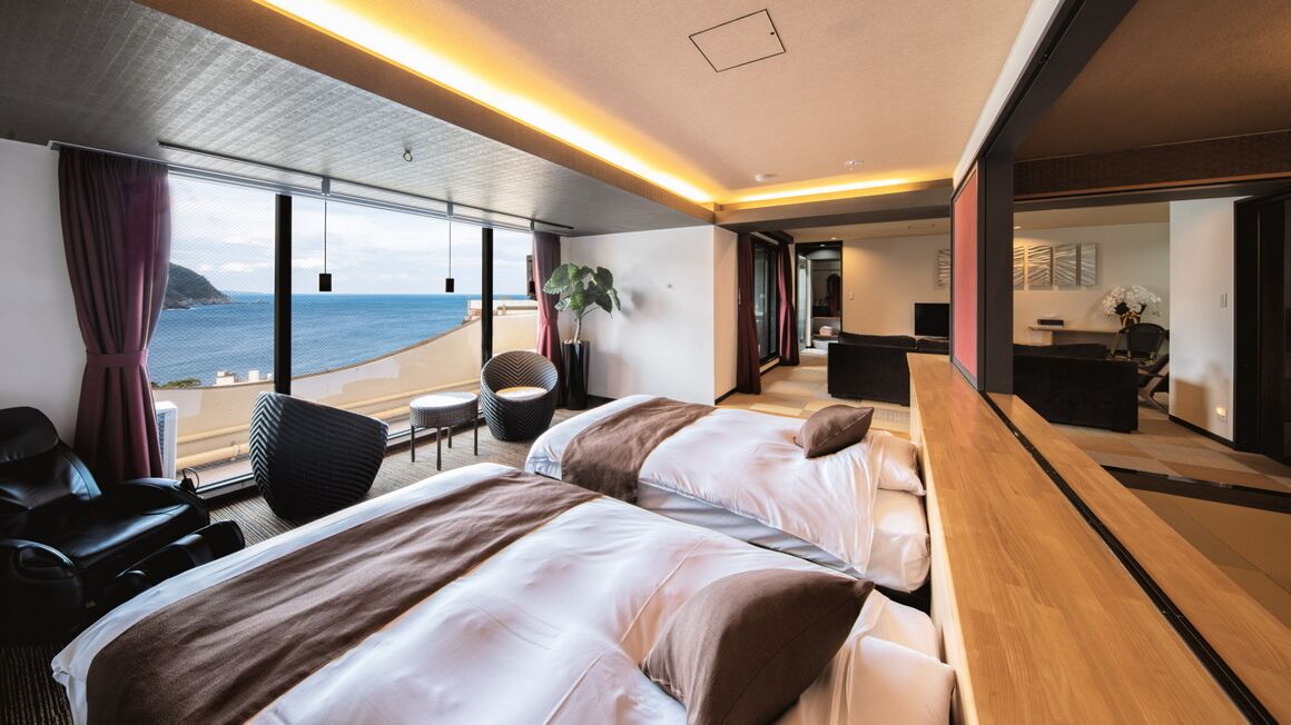 A modern hotel room with two beds, a wooden desk, a view of the sea, and a balcony.
