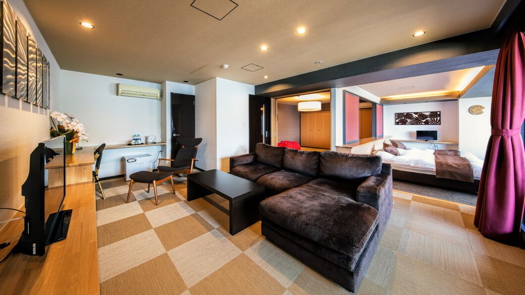 A modern hotel room with a sofa, a coffee table, a desk with a chair, a bed with a red blanket, and a TV.