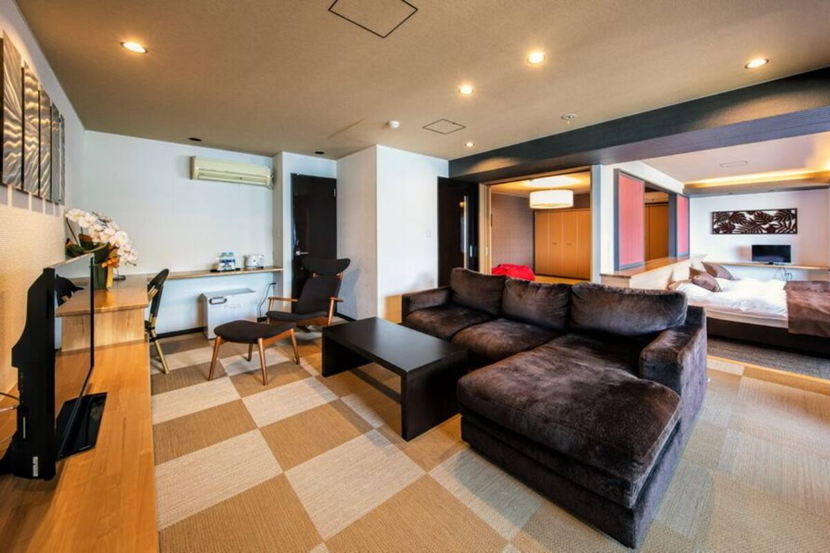 A modern hotel room with a sofa, a coffee table, a desk with a chair, a bed with a red blanket, and a TV.