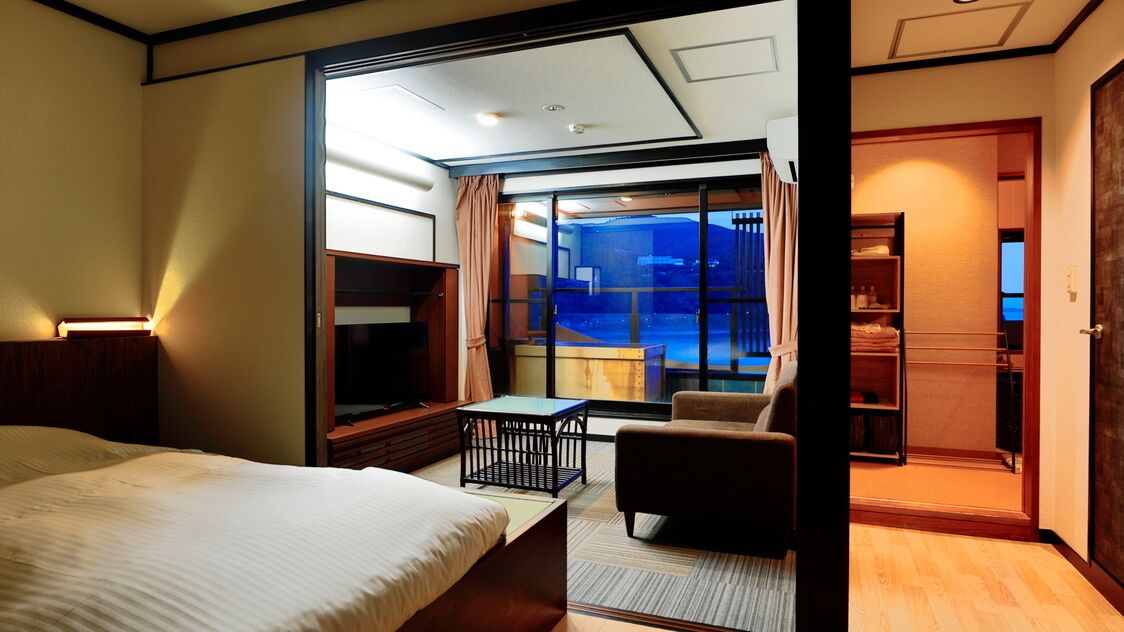 A hotel room with a bed, a small table, a chair, a TV, and a view of the outside.