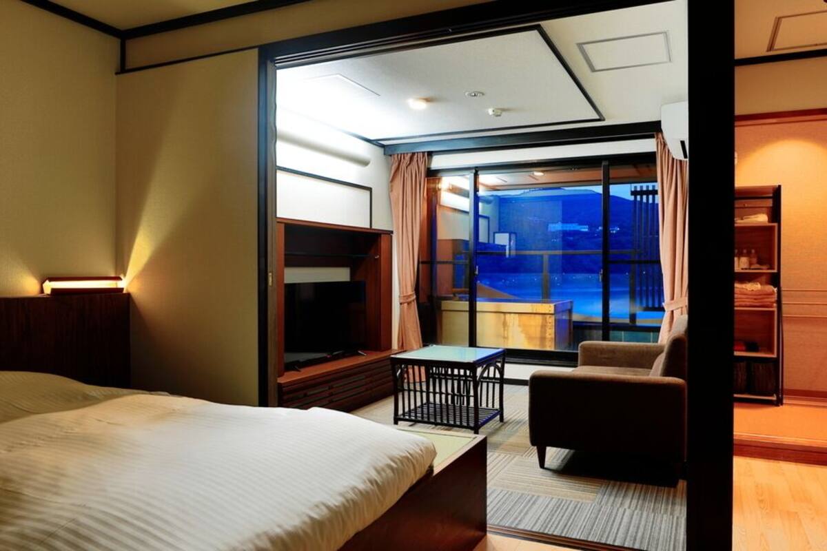 A hotel room with a bed, a small table, a chair, a TV, and a view of the outside.