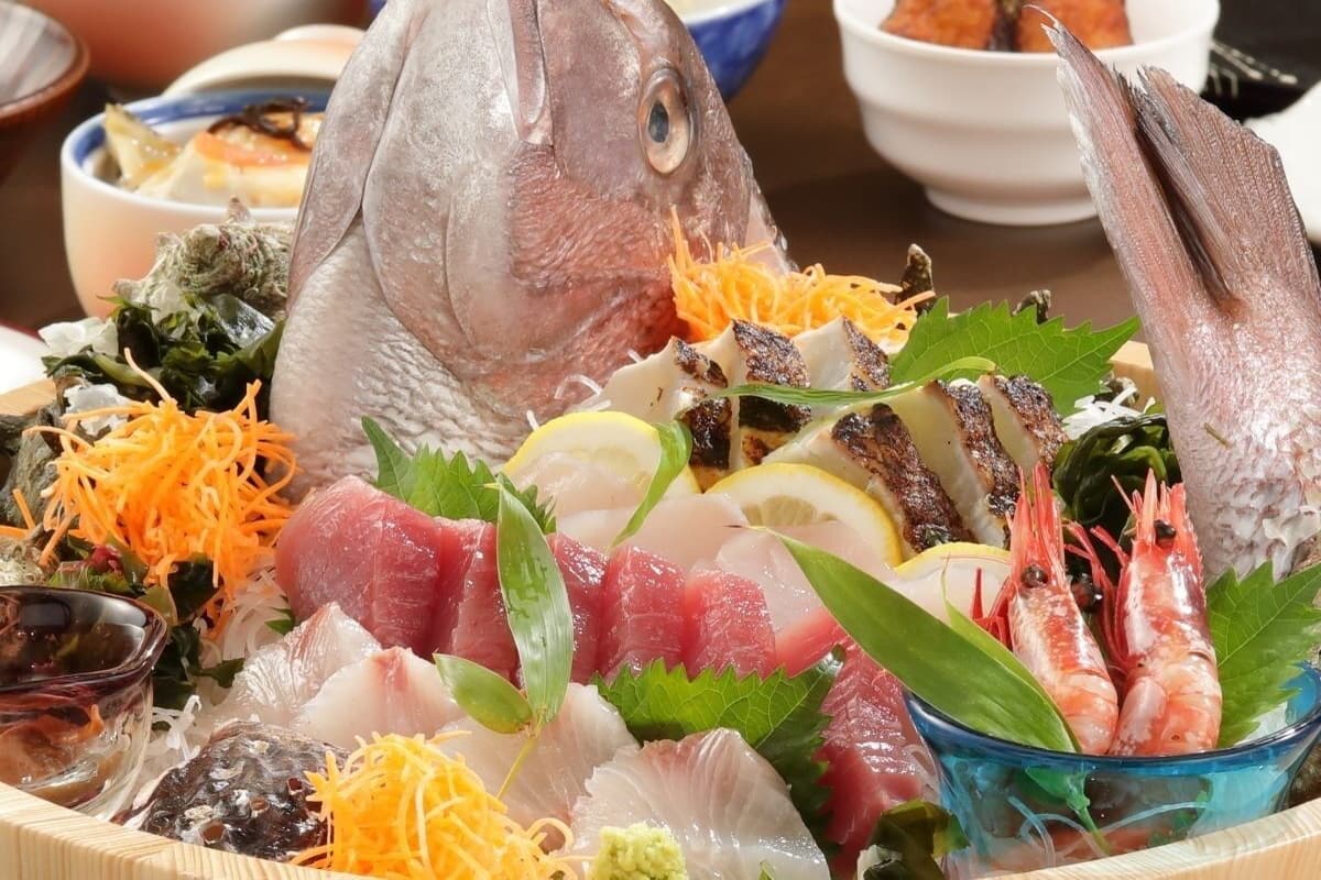 A variety of seafood including fish, shrimp, and possibly sashimi, arranged on a table with a bowl of soup and other dishes in the background.